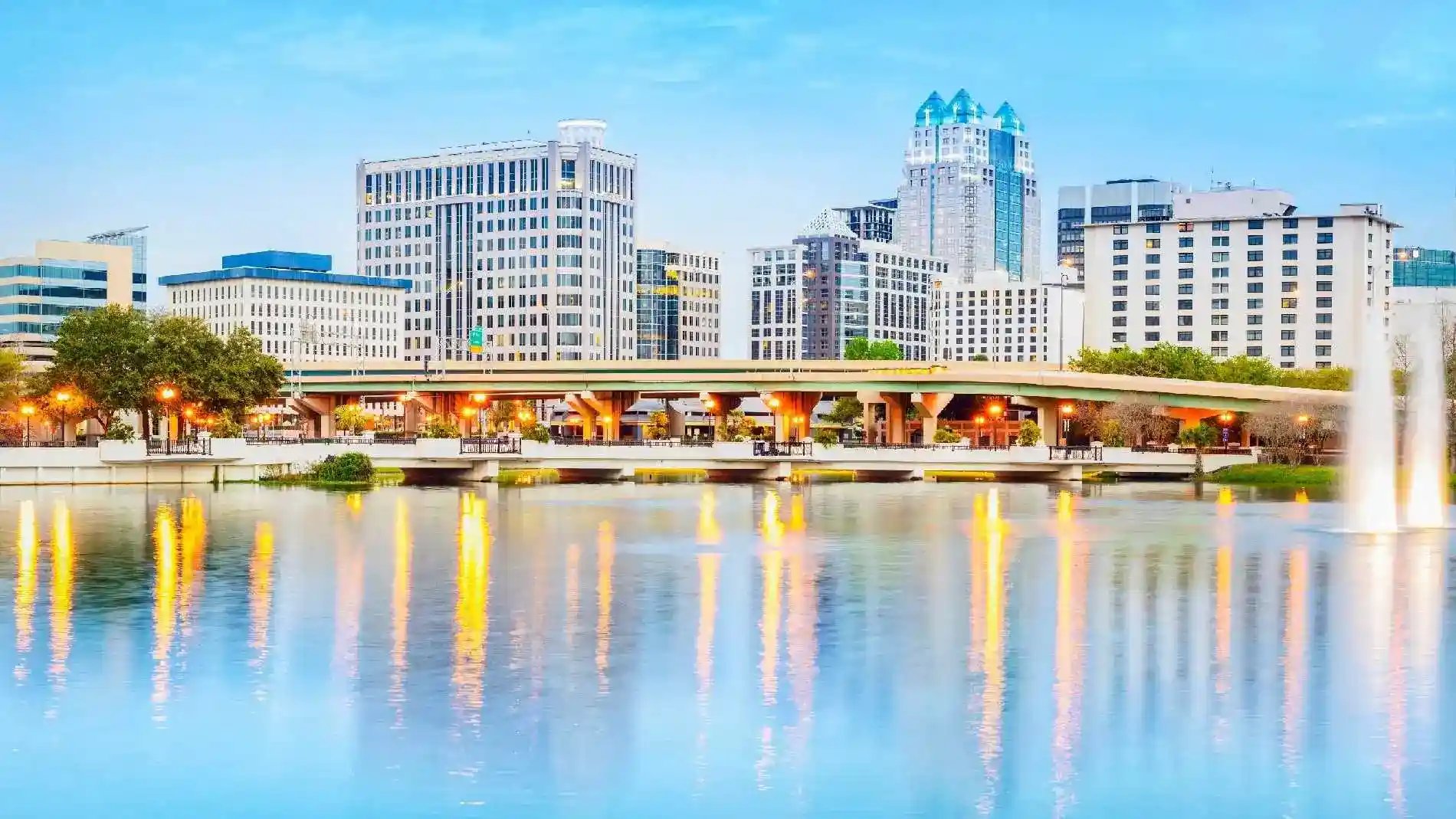 Orlando city skyline with tall buildings reflecting on the calm waters of Lake Eola, surrounded by lush greenery and a clear blue sky